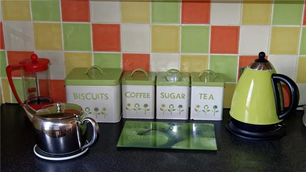 На кухне стоит зеленый электрочайник, металлический чайник для заваривания чая, стеклянный заварочный чайник, четрые емкости для сыпучих веществ с надписями на английском языке